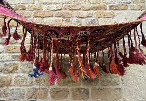 fig 3. A handwoven red wool tribal textile featuring geometric motifs, hanging tassels, leather loops, and top and bottom wooden support rods.