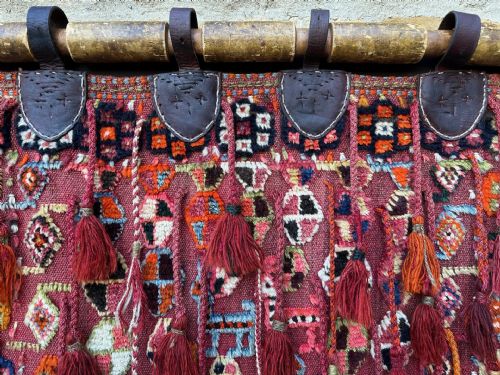 fig 4. A handwoven red wool tribal textile featuring geometric motifs, hanging tassels, leather loops, and top and bottom wooden support rods.