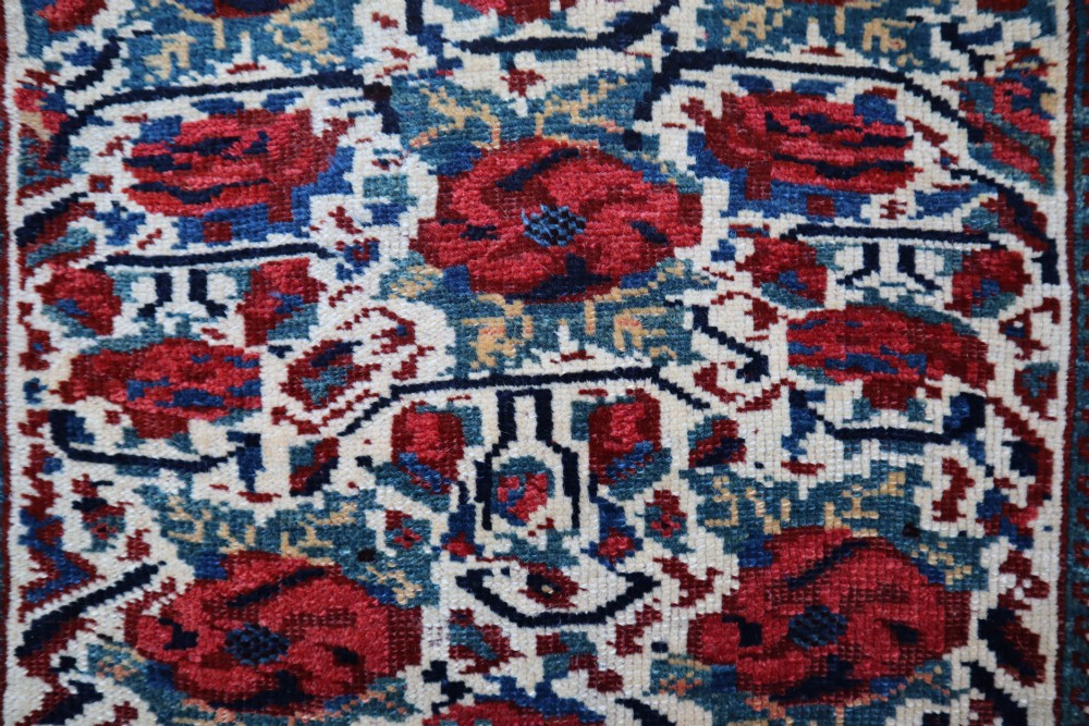 A square antique woven wool saddlebag face featuring stylised red floral rosettes on an ivory ground with an indigo blue geometric border and braided loops along the top edge. View II