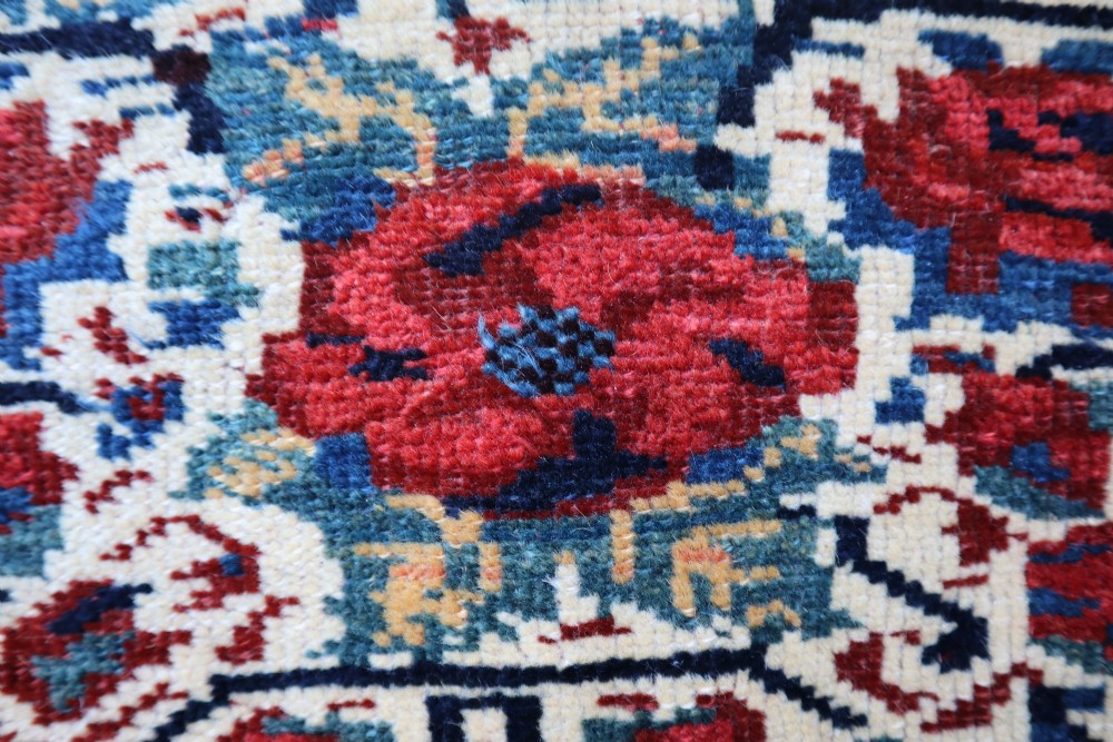 A square antique woven wool saddlebag face featuring stylised red floral rosettes on an ivory ground with an indigo blue geometric border and braided loops along the top edge. View III