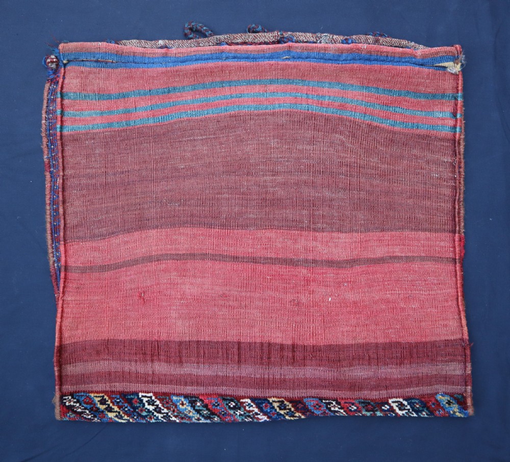 A square antique woven wool saddlebag face featuring stylised red floral rosettes on an ivory ground with an indigo blue geometric border and braided loops along the top edge. View IV