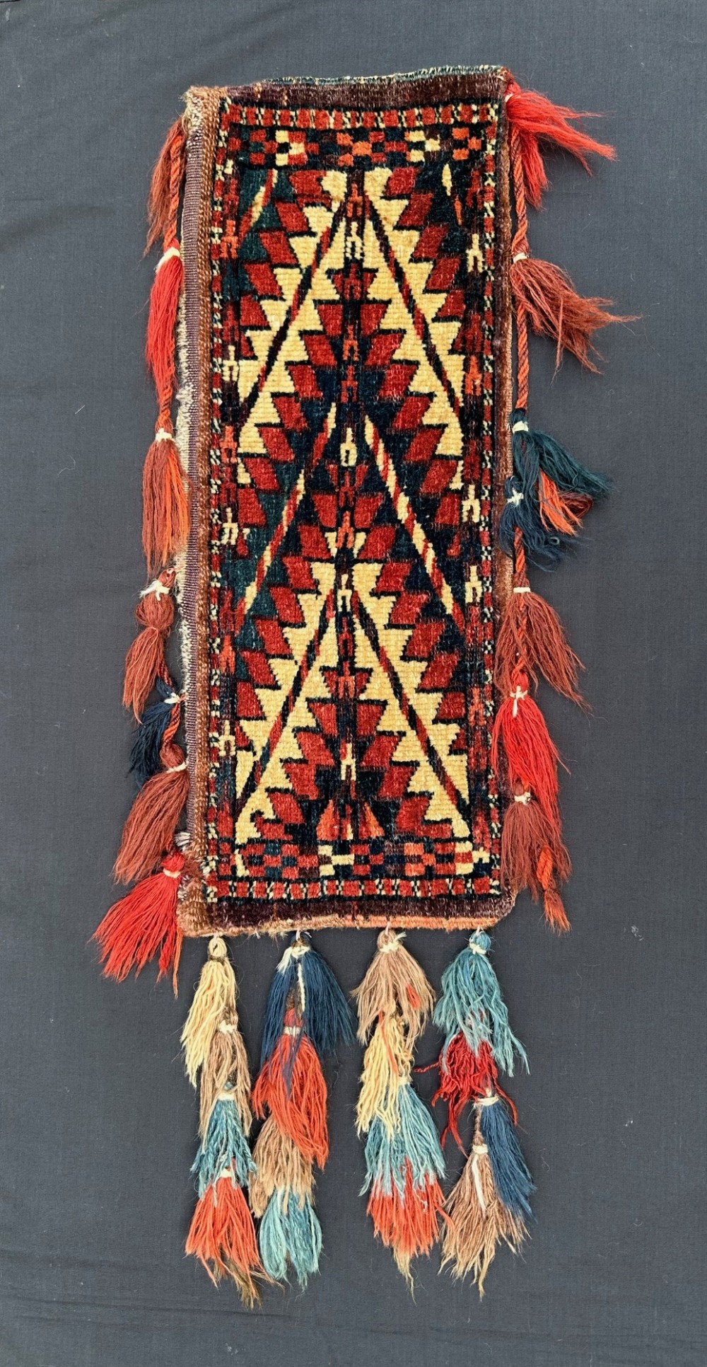 A narrow, elongated hand-woven Turkmen wool spindle bag with geometric serrated diamond patterns in red, navy blue, and cream, featuring long wool tassels along the sides and base.