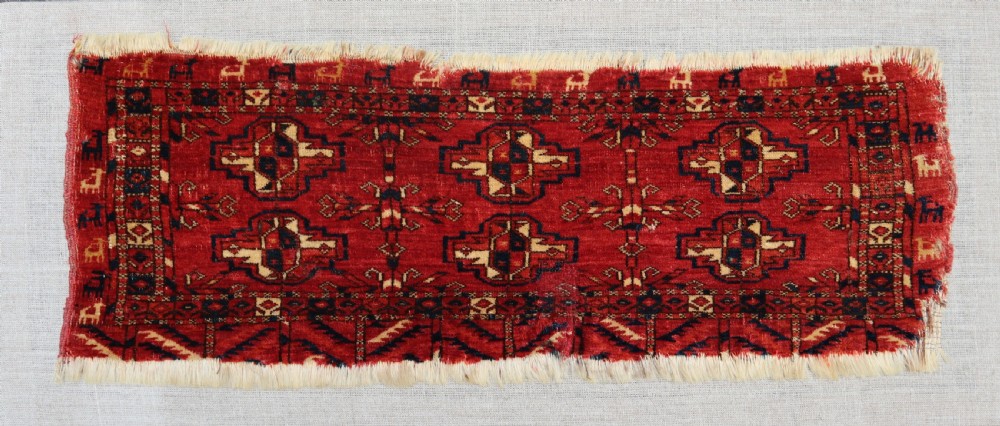 An antique hand-woven red wool Teke Turkmen rug fragment with dark navy geometric gols and small animal figures mounted on a neutral linen backing board. View II