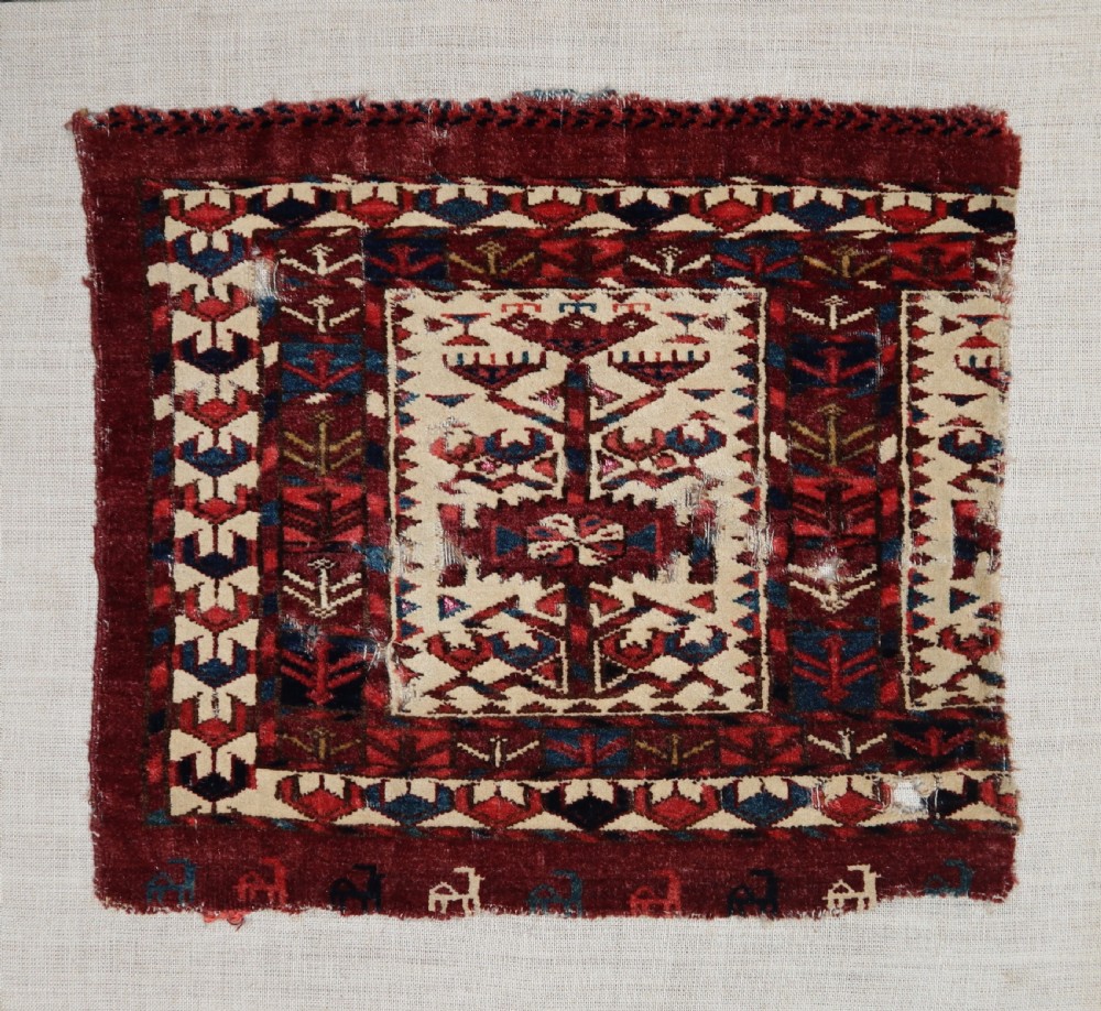 A hand-woven wool Turkmen Teke mafrash textile fragment featuring geometric motifs in deep red, blue, and cream, mounted on a light beige backing. View II