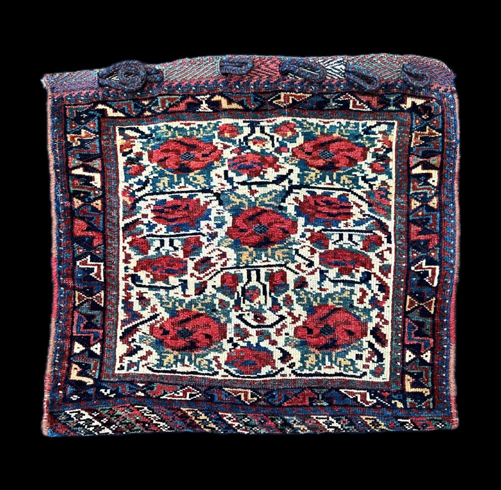 A square antique woven wool saddlebag face featuring stylised red floral rosettes on an ivory ground with an indigo blue geometric border and braided loops along the top edge.