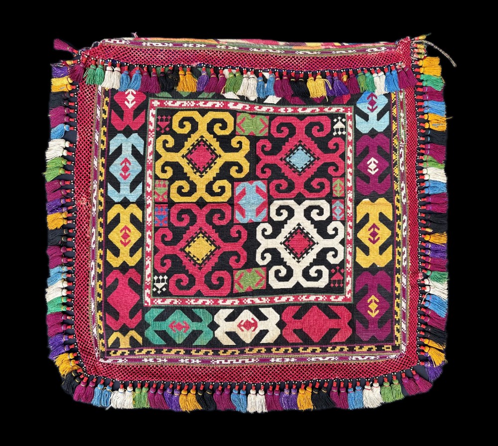 A square antique Central Asian Lakai embroidered textile featuring bold geometric scroll motifs in yellow, magenta, white, and blue surrounded by multi-coloured tassel fringe.