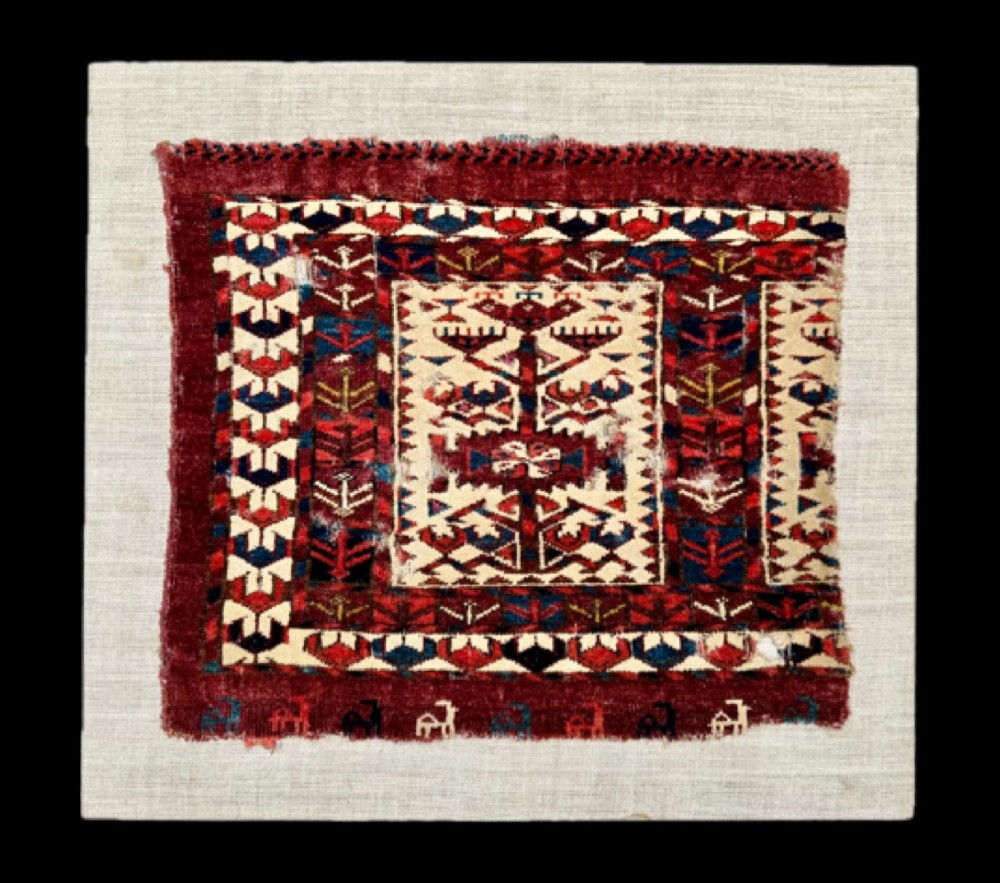 A hand-woven wool Turkmen Teke mafrash textile fragment featuring geometric motifs in deep red, blue, and cream, mounted on a light beige backing.
