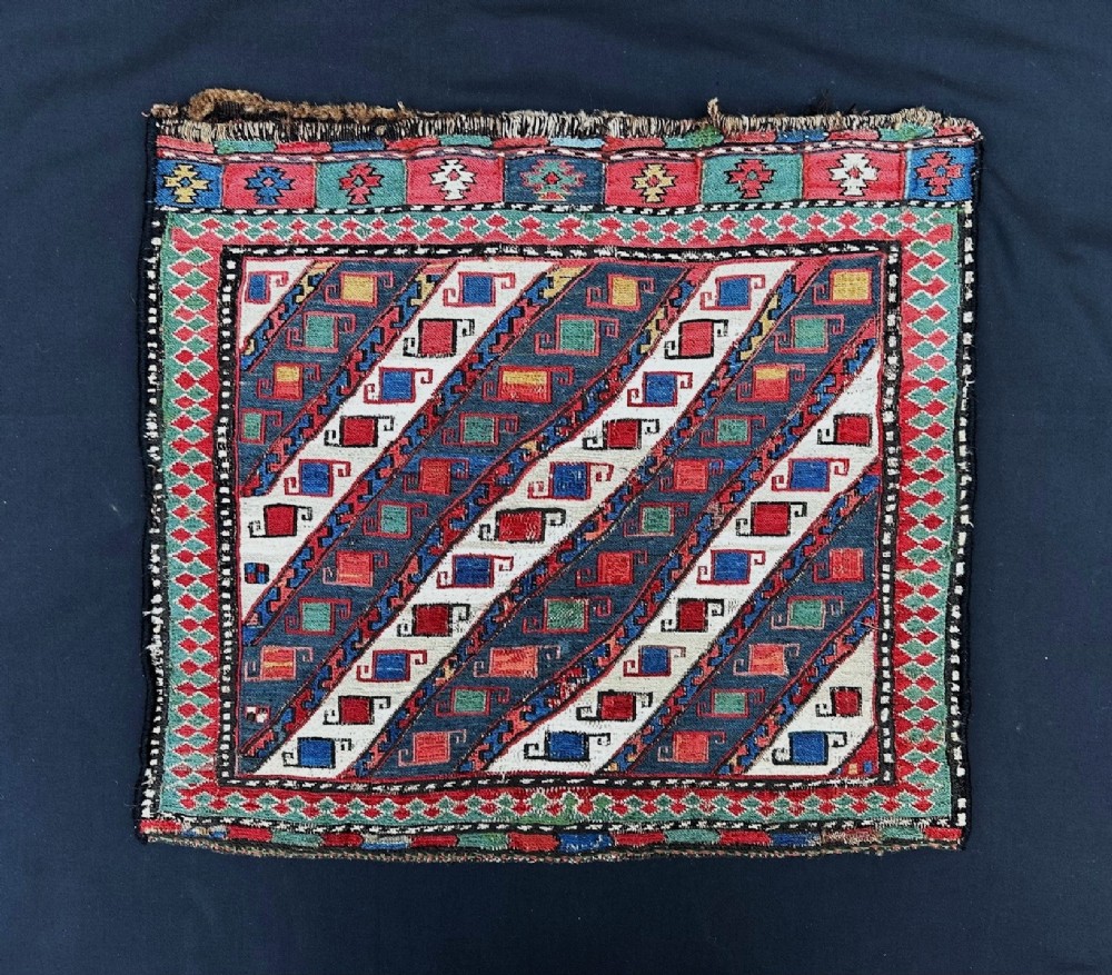 A square handwoven Shahsevan wool saddlebag face featuring diagonal blue and white stripes filled with red geometric hooked motifs within patterned borders.