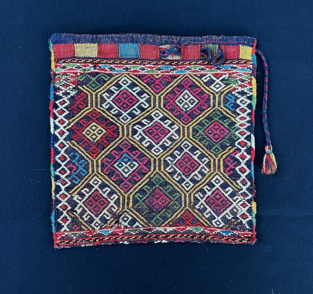 A square flatweave textile bag featuring geometric diamond motifs in red, yellow, green, magenta, and white wool against a dark background with a braided cord.