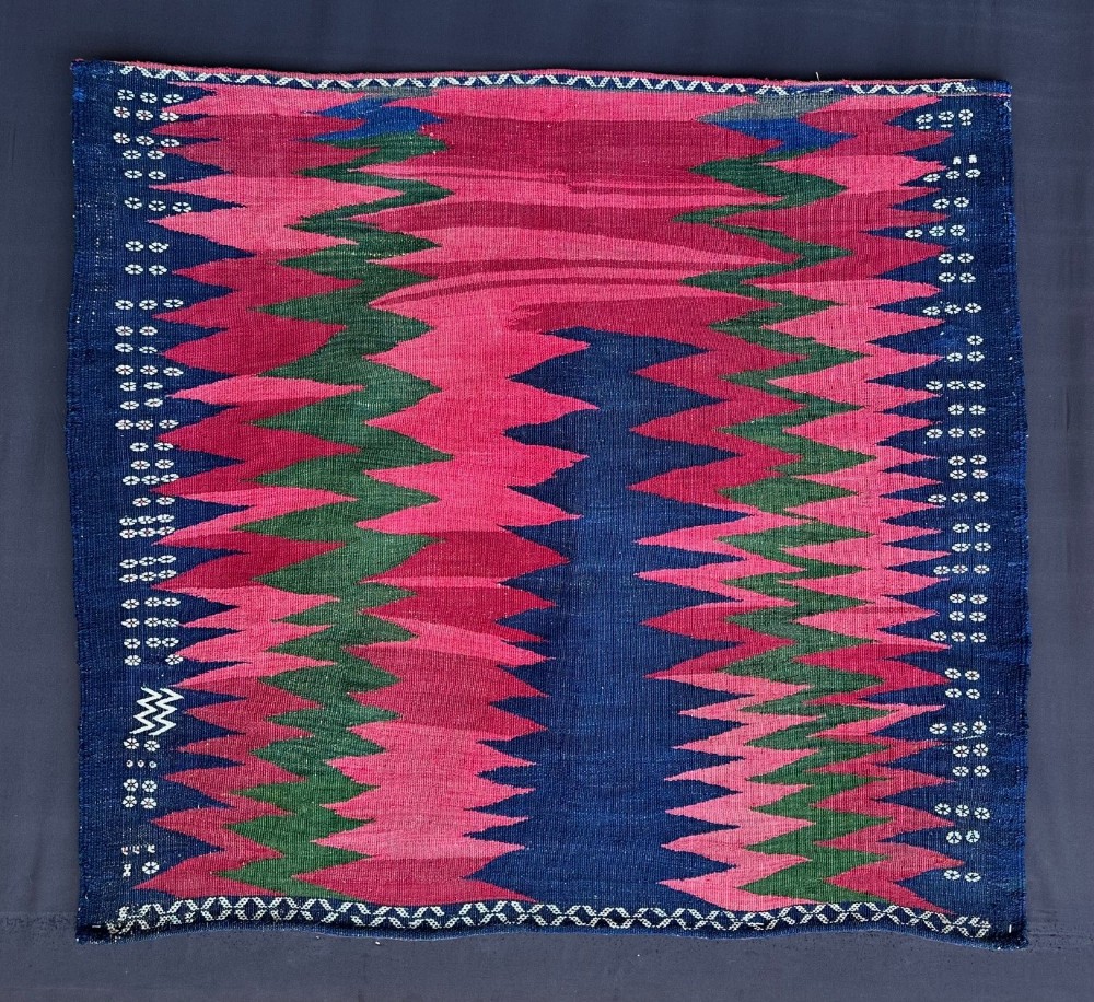 A square handwoven flatweave textile featuring bold zigzag patterns in magenta, green, and dark blue wool with decorative border motifs.