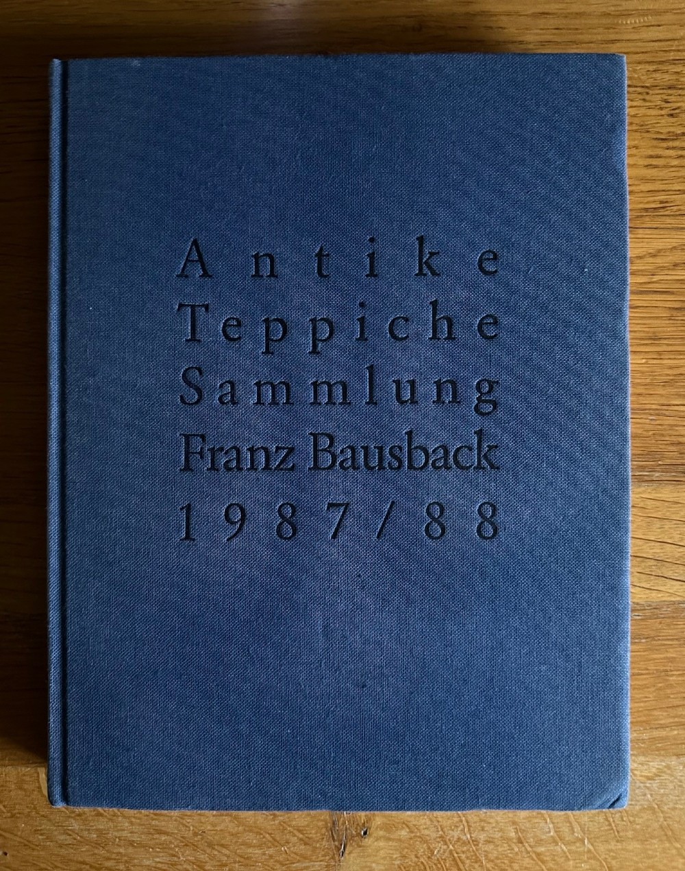 A navy blue cloth-bound hardback book featuring printed black text reading Antike Teppiche Sammlung Franz Bausback 1987/88.