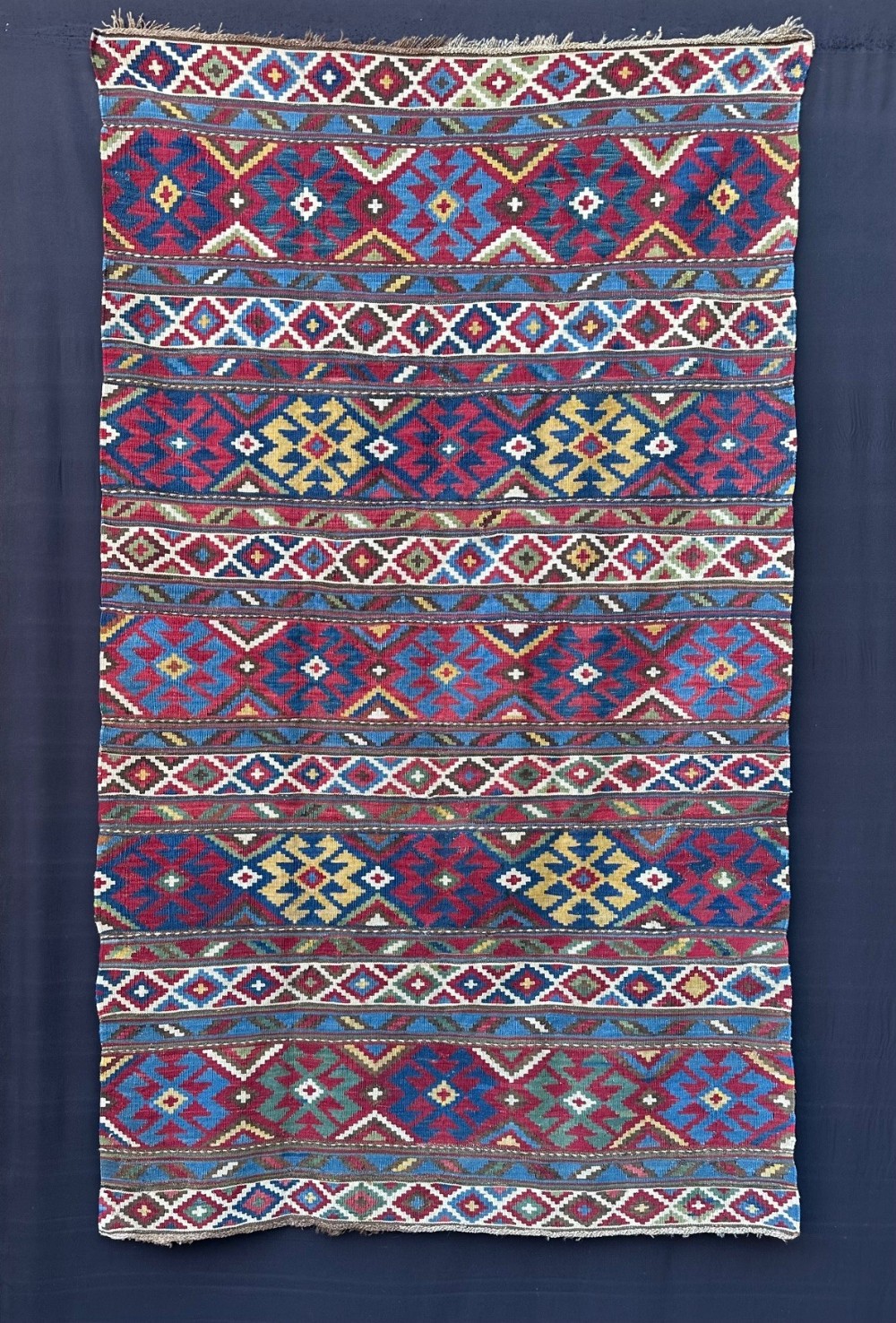 An antique handwoven wool Shirvan kilim rug featuring horizontal bands of geometric diamond and star motifs in shades of red, blue, yellow, and ivory.