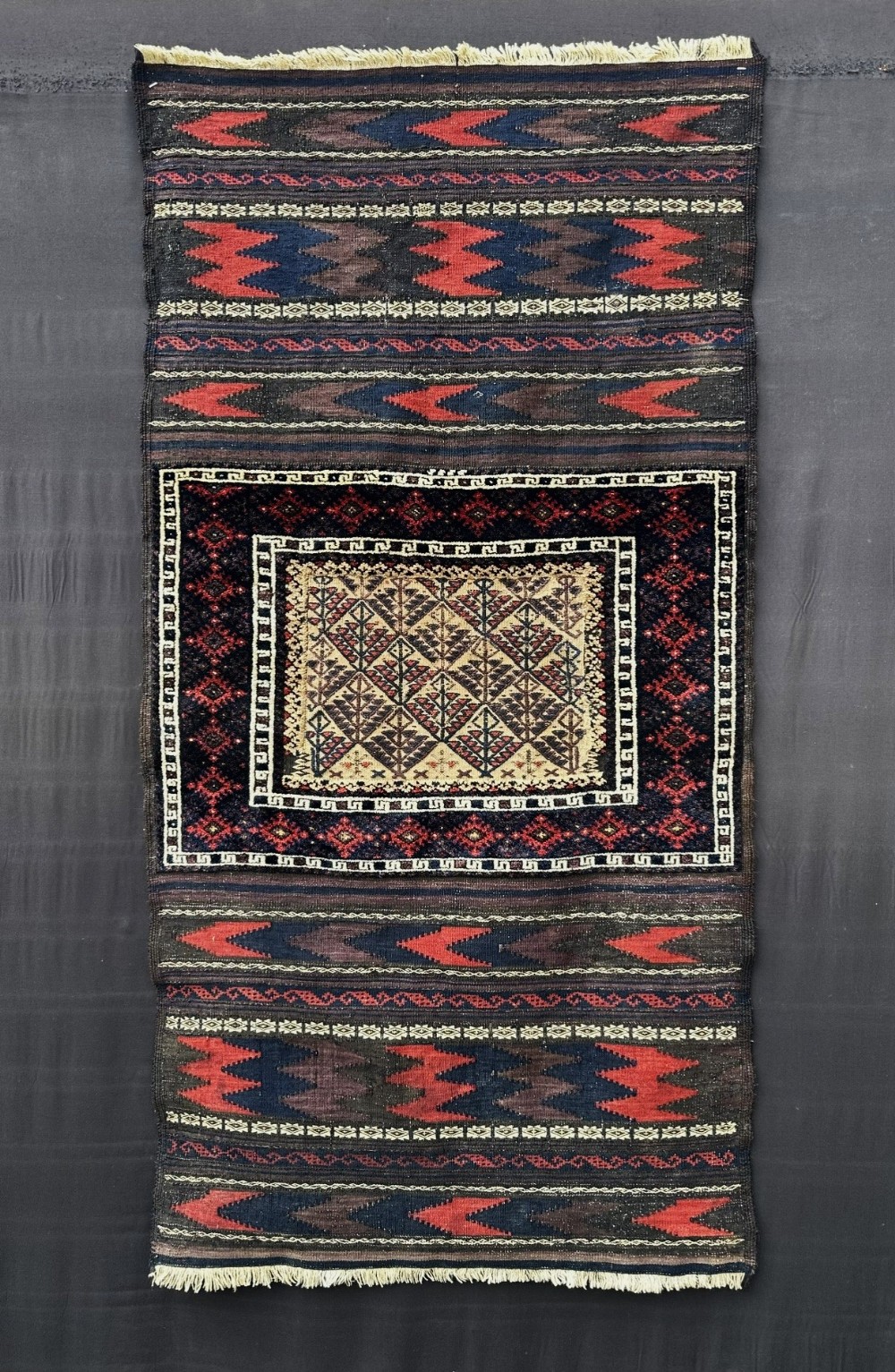 An antique handwoven Baluch Sofreh flatweave textile featuring geometric chevron bands in dark red and navy framing a central panel with tree motifs.