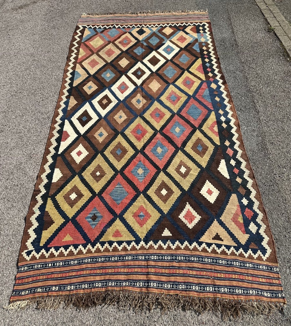 An antique handwoven Persian Qashqai wool kilim rug featuring repeating geometric diamond motifs in terracotta, dark brown, blue, yellow, and cream hues.