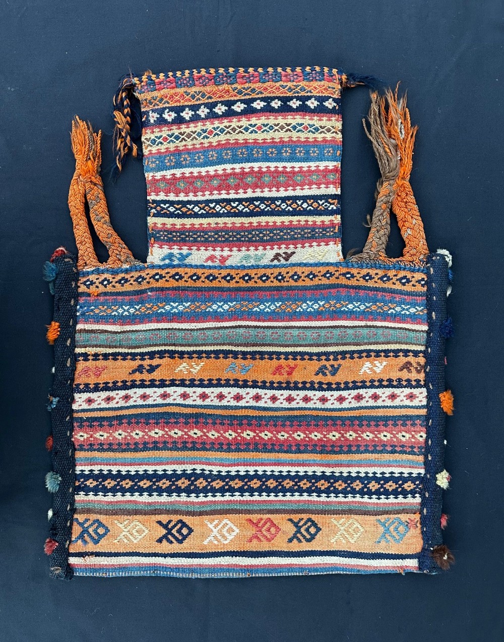 A handwoven antique Qashqai salt bag featuring horizontal bands of geometric patterns in orange, blue, red, and white wool with braided side loops.