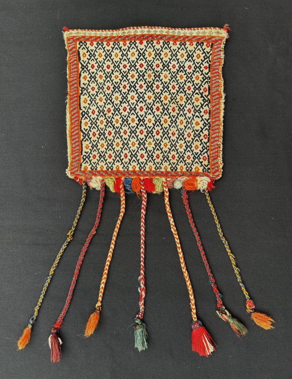 A square woven Persian tribal vanity bag featuring a black and red geometric diamond pattern on a cream background, finished with woven borders and long braided tassels ending in colourful fringes.