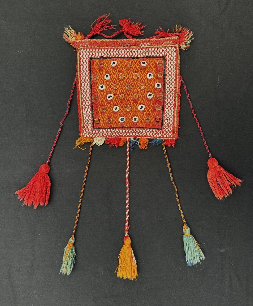 A small square woven wool Qashqai vanity bag featuring a red geometric patterned centre, braided borders, and multi-coloured hanging tassels.