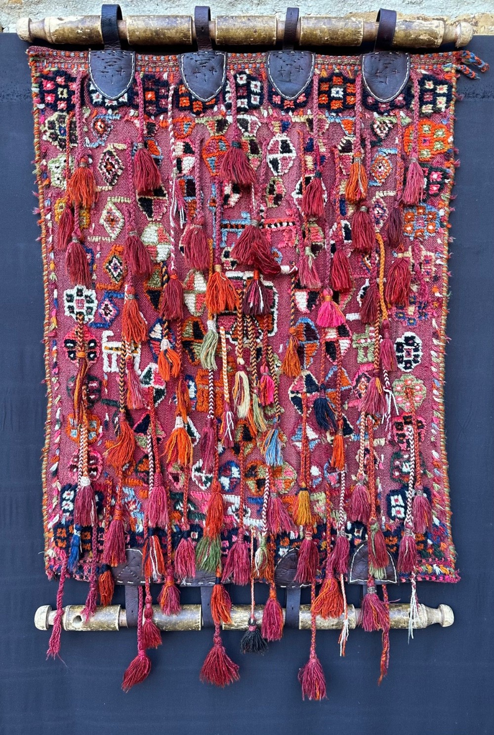A handwoven red wool tribal textile featuring geometric motifs, hanging tassels, leather loops, and top and bottom wooden support rods.
