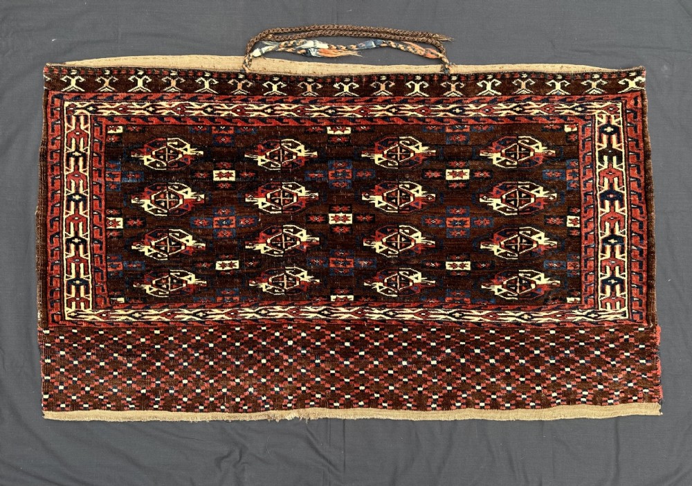 Handwoven antique Turkmen Yomut chuval featuring repeating tribal gul motifs in deep red and brown tones with a patterned lower elem and braided top handles.