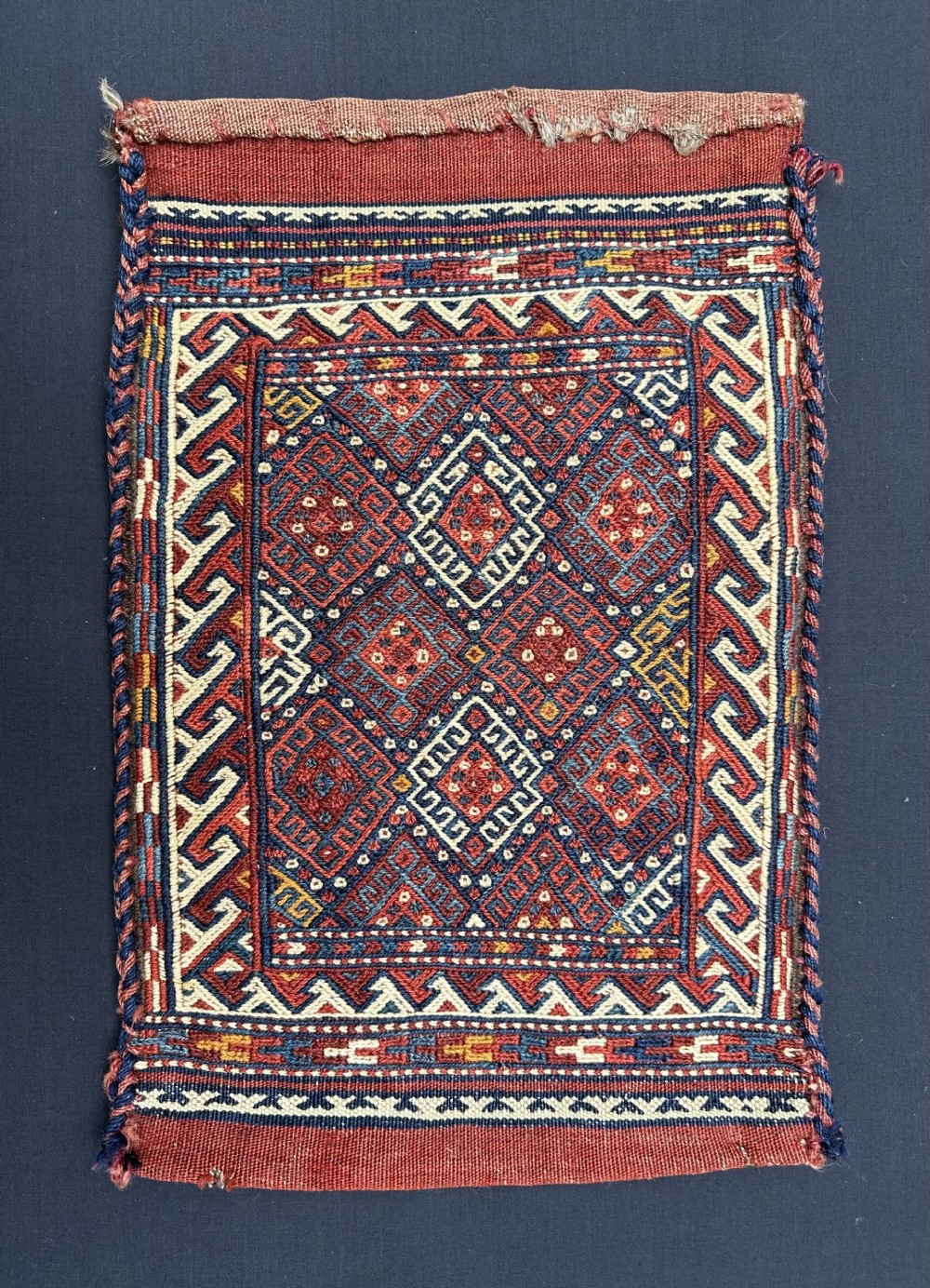 A rectangular hand-woven tribal wool bag face with geometric diamond motifs in terracotta red, indigo blue, and cream.