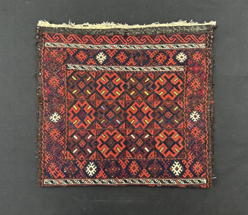 A square antique handwoven Baluch tribal bag face featuring geometric hooked motifs in dark red, navy blue, yellow, and white wool.