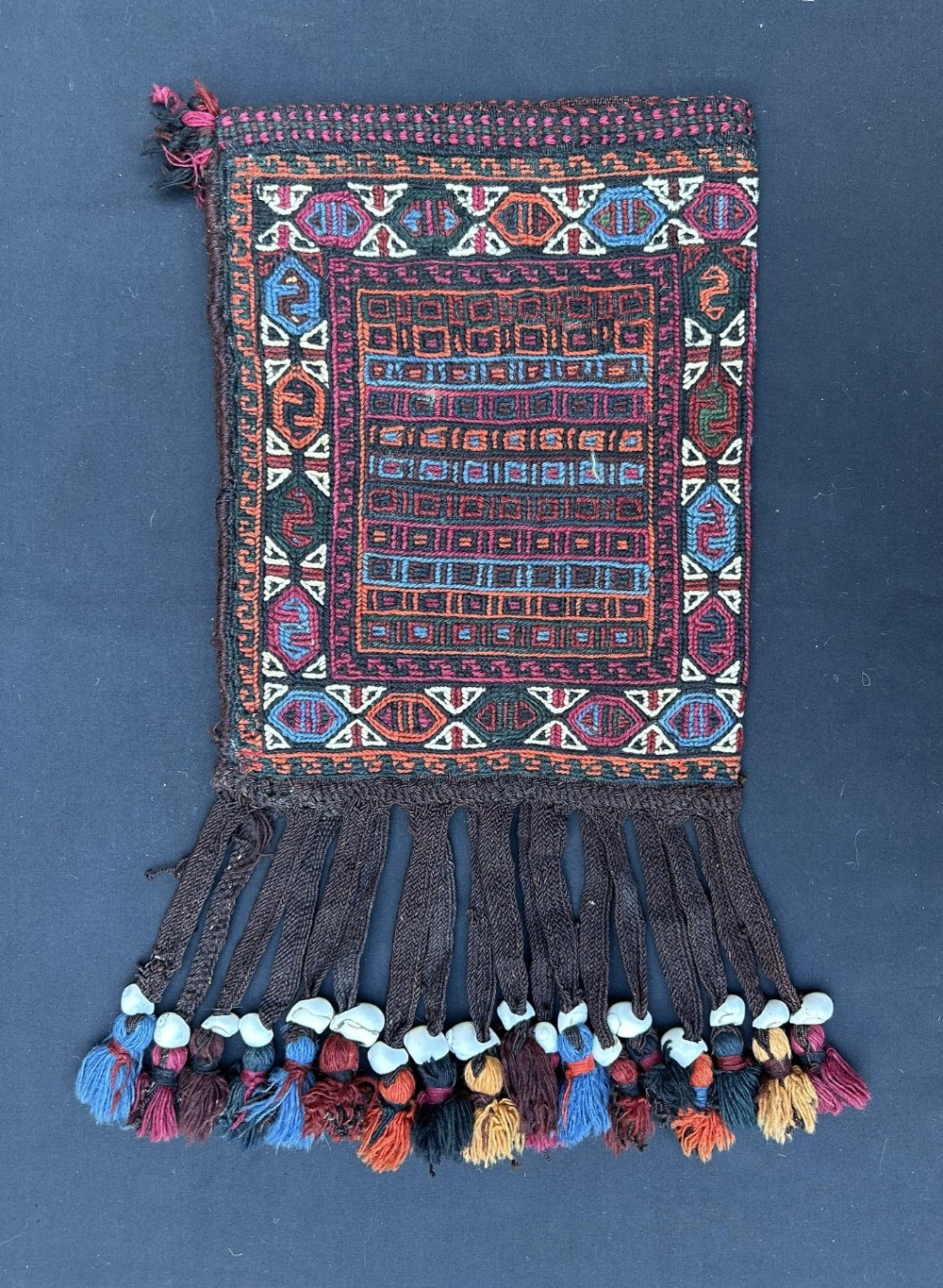 An antique Persian tribal bag displaying geometric woven patterns in red, blue, and orange with a heavy fringe embellished with cowrie shells and tassels.
