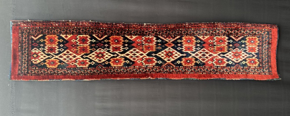 A long, narrow handwoven wool textile featuring intricate geometric diamond motifs in deep red, dark navy, and ivory shades.