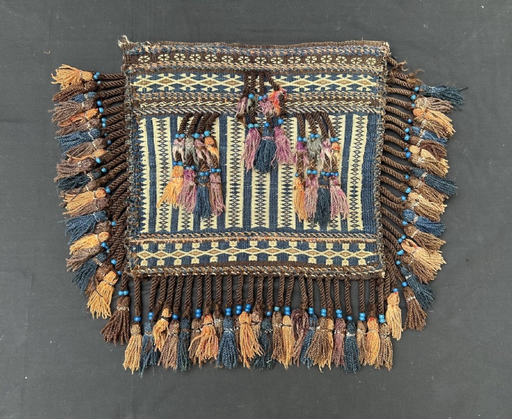 A handwoven square Baluch tribal storage bag face featuring dark blue and beige geometric flatweave patterns, decorated with braided tassels and small blue beads along the edges.