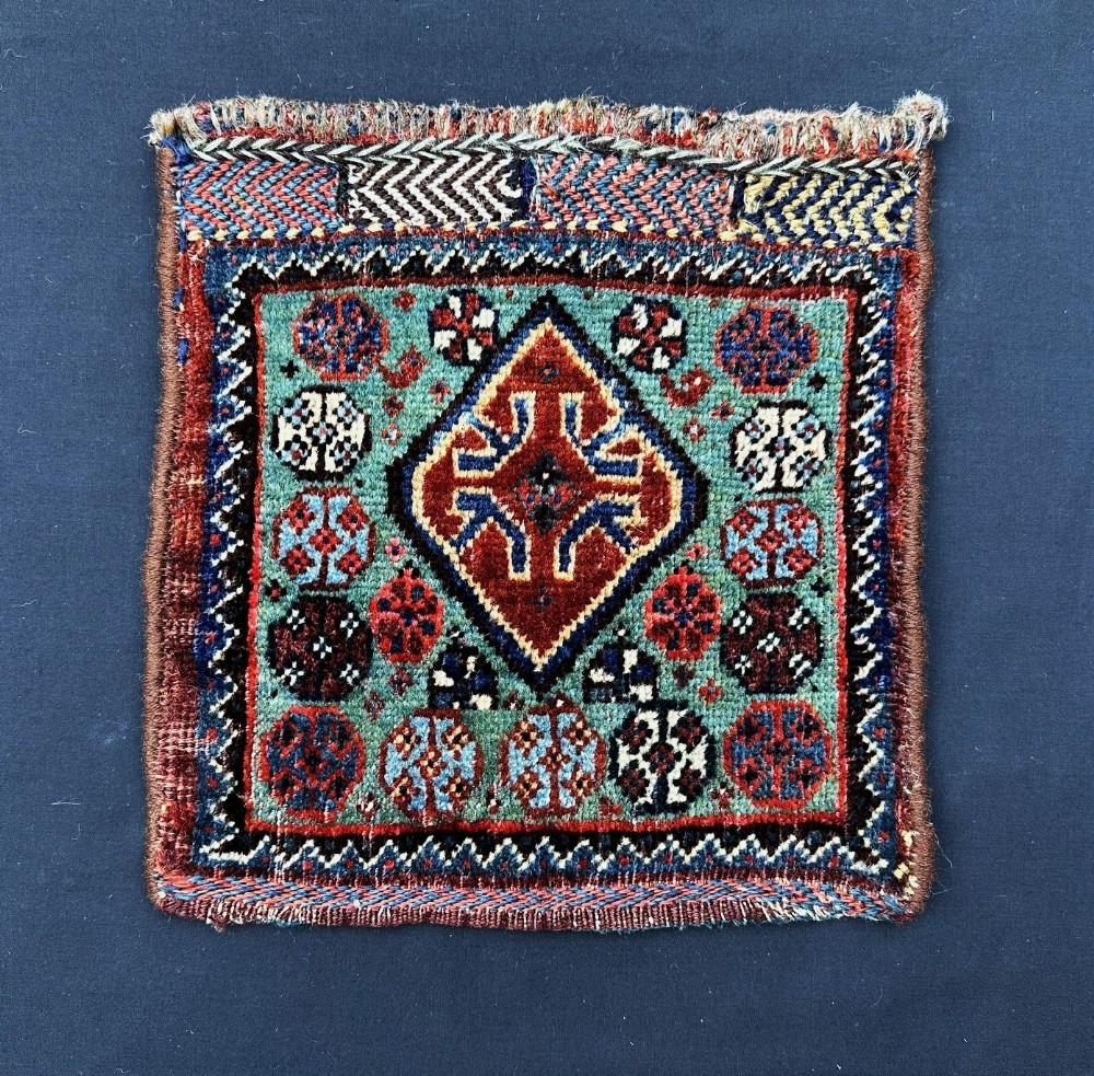 A square antique Persian Khamseh tribal wool bagface featuring a central diamond medallion surrounded by geometric rosettes on a turquoise ground with woven patterned borders.
