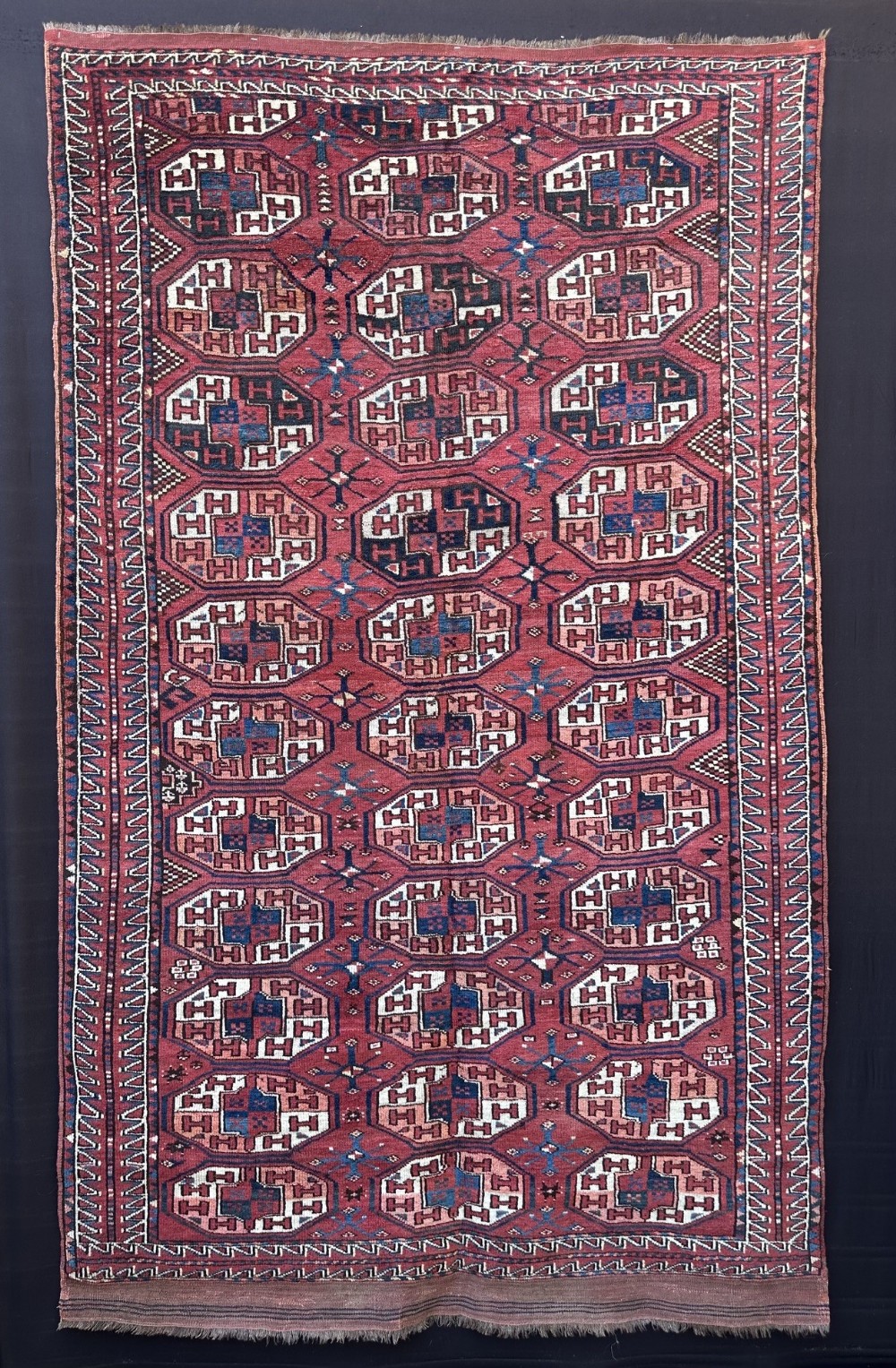 An antique hand-knotted Turkmen wool carpet featuring repeating octagonal tribal gul motifs in shades of deep red, navy blue, ivory, and terracotta within geometric borders.