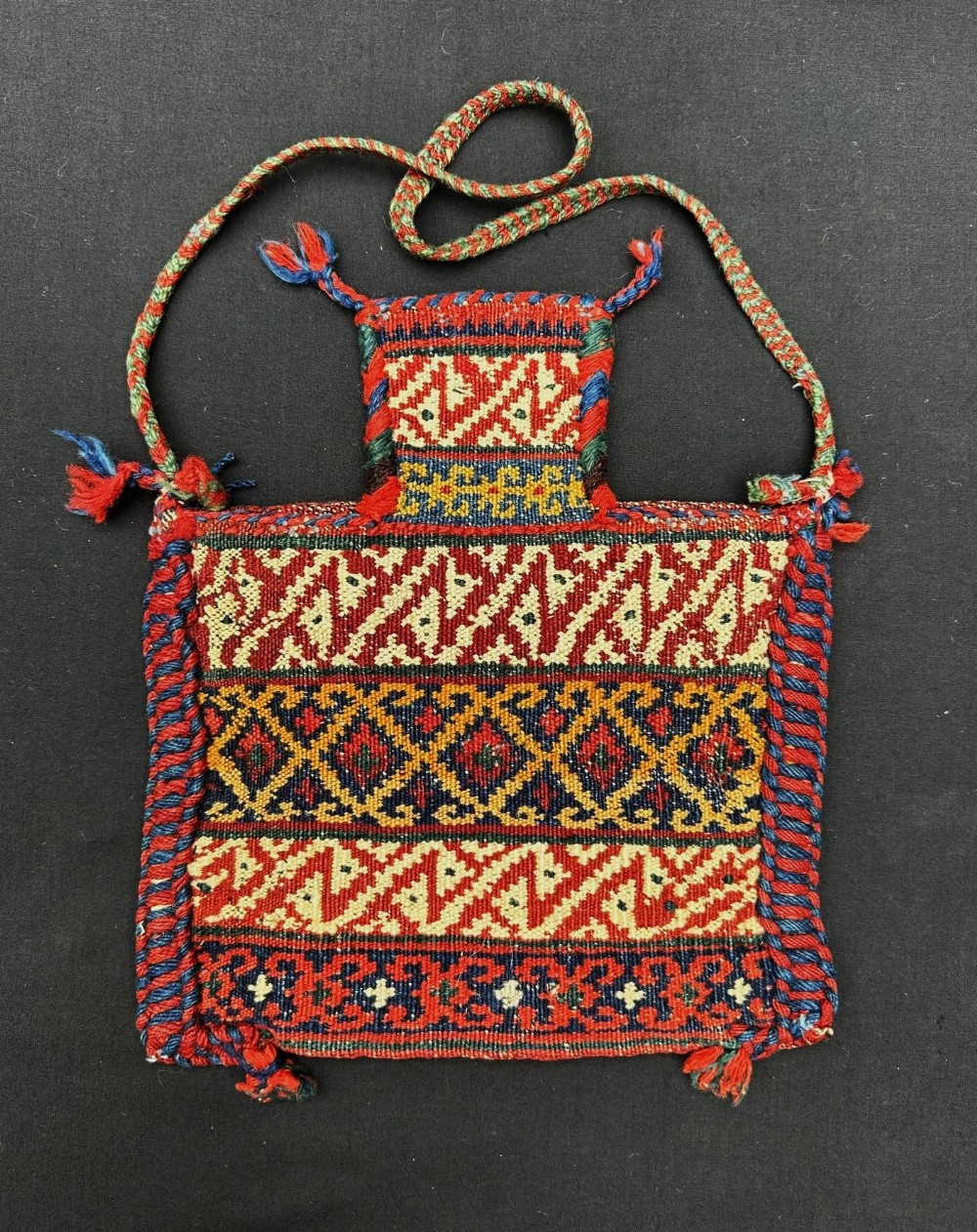 A small hand-woven wool tribal pouch featuring geometric patterns in red, cream, blue, and yellow with a narrow neck opening and braided shoulder strap.