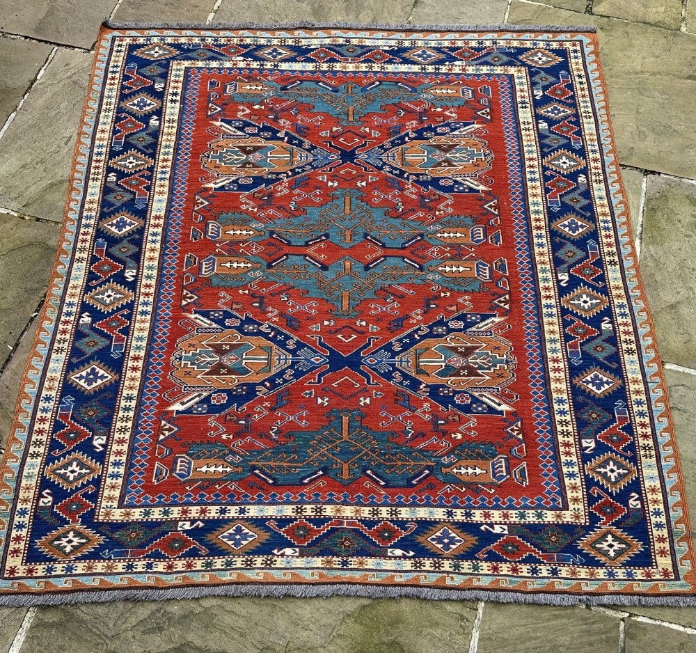 An antique Daghestan Soumak flatweave wool rug featuring central geometric blue medallions framed by a terracotta red field and multi-layered geometric borders.