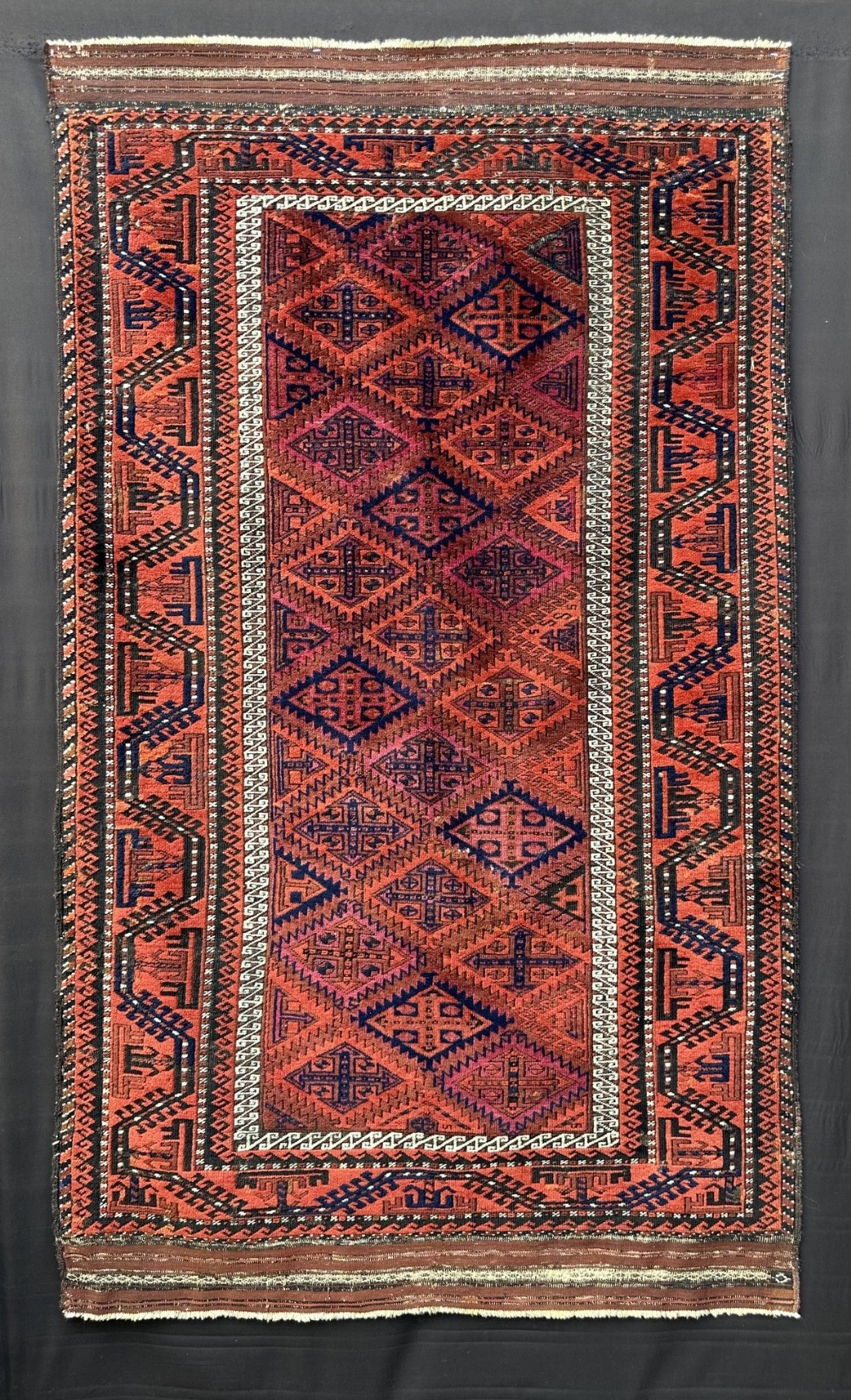 A rectangular hand-knotted wool rug featuring repeating geometric diamond latch-hook patterns in terracotta red, deep blue, and ivory accents.