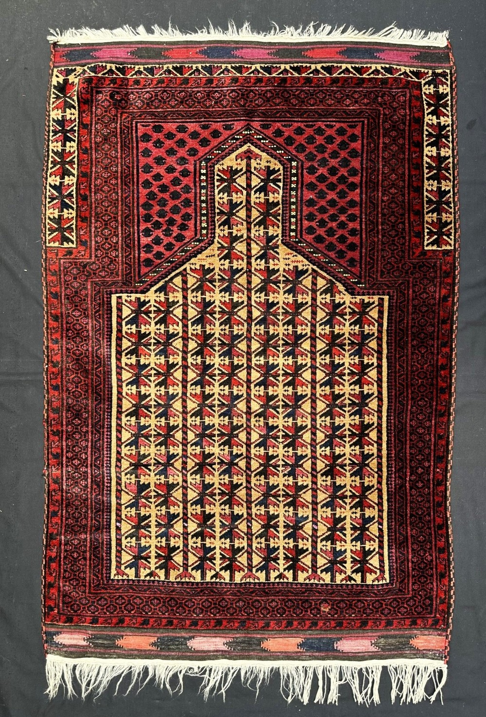 A hand-knotted rectangular wool prayer rug featuring a dark red and beige geometric mihrab design with tribal motifs and fringed ends.
