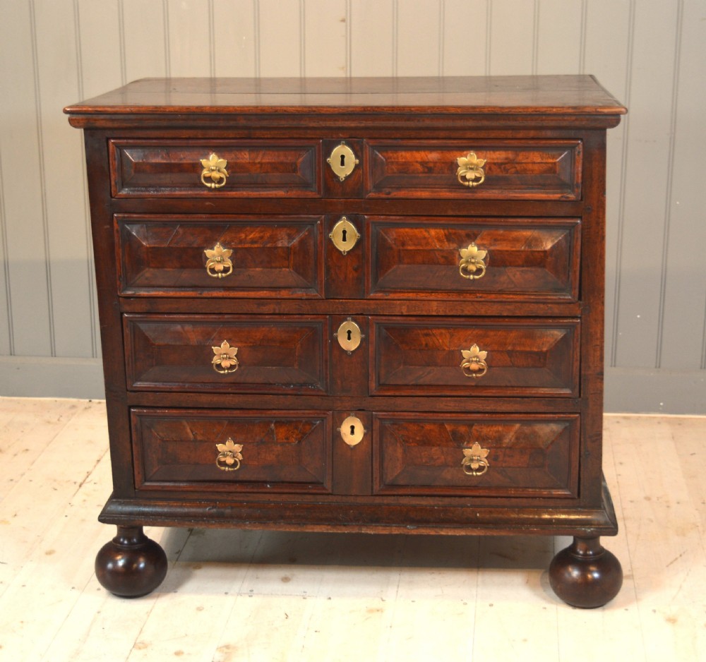 A 17th-century walnut chest of drawers with four moulded drawer fronts, brass drop handles, brass keyhole escutcheons, and large turned bun feet.