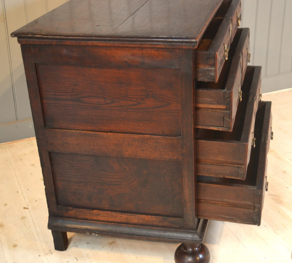 A 17th-century walnut chest of drawers with four moulded drawer fronts, brass drop handles, brass keyhole escutcheons, and large turned bun feet. View VI