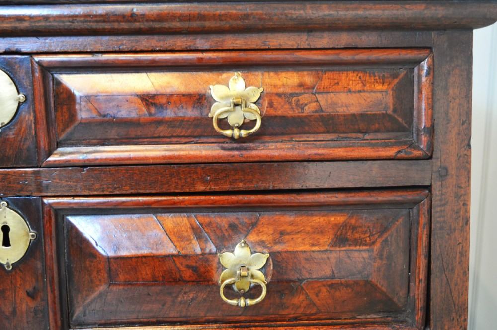 A 17th-century walnut chest of drawers with four moulded drawer fronts, brass drop handles, brass keyhole escutcheons, and large turned bun feet. View VII
