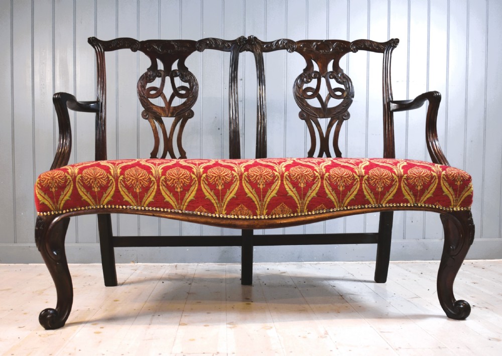 An 18th-century mahogany double-chair-back settee featuring pierced carved back splats, red patterned fabric upholstery, cabriole front legs, and curved armrests.
