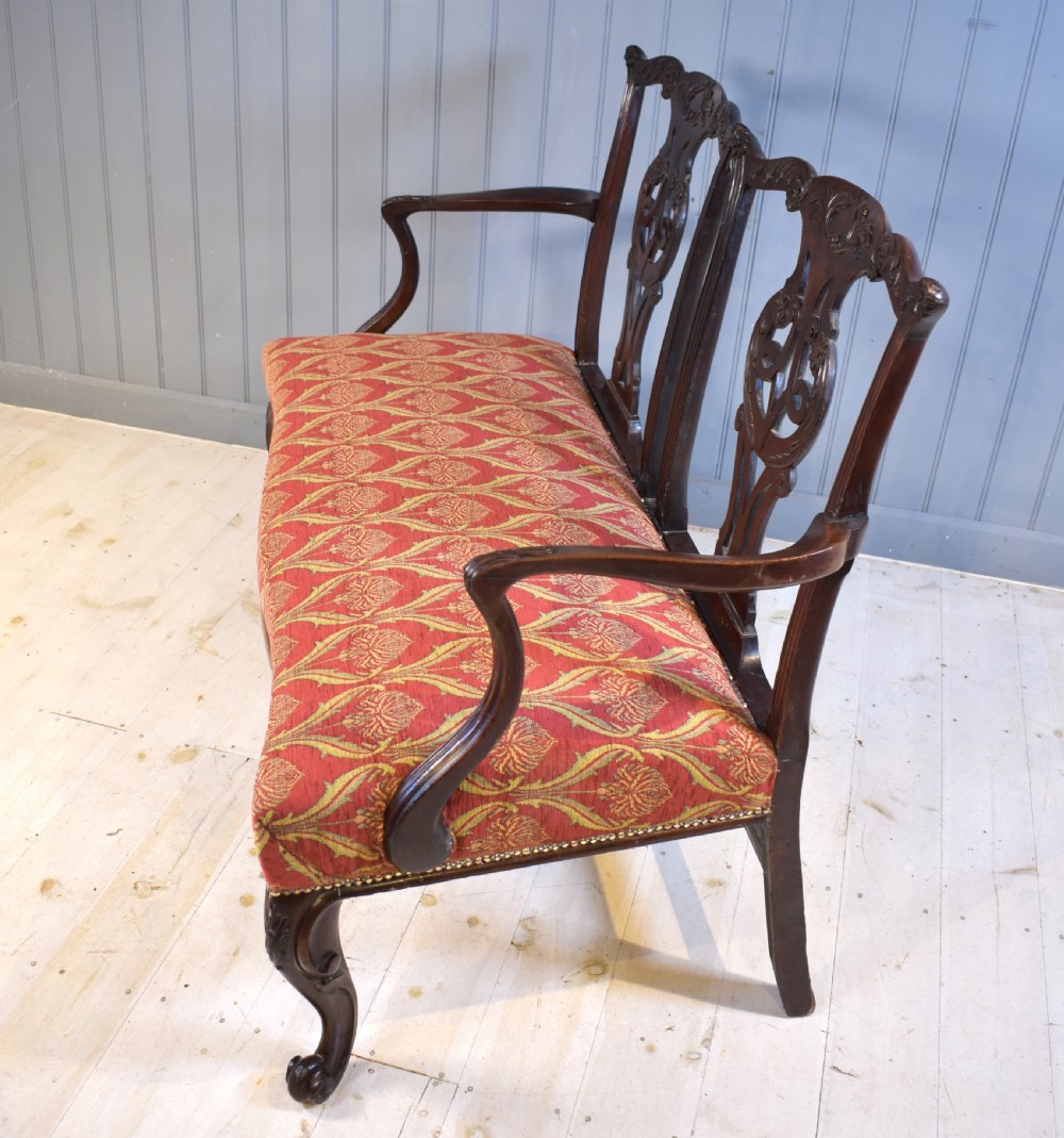 An 18th-century mahogany double-chair-back settee featuring pierced carved back splats, red patterned fabric upholstery, cabriole front legs, and curved armrests. View VI