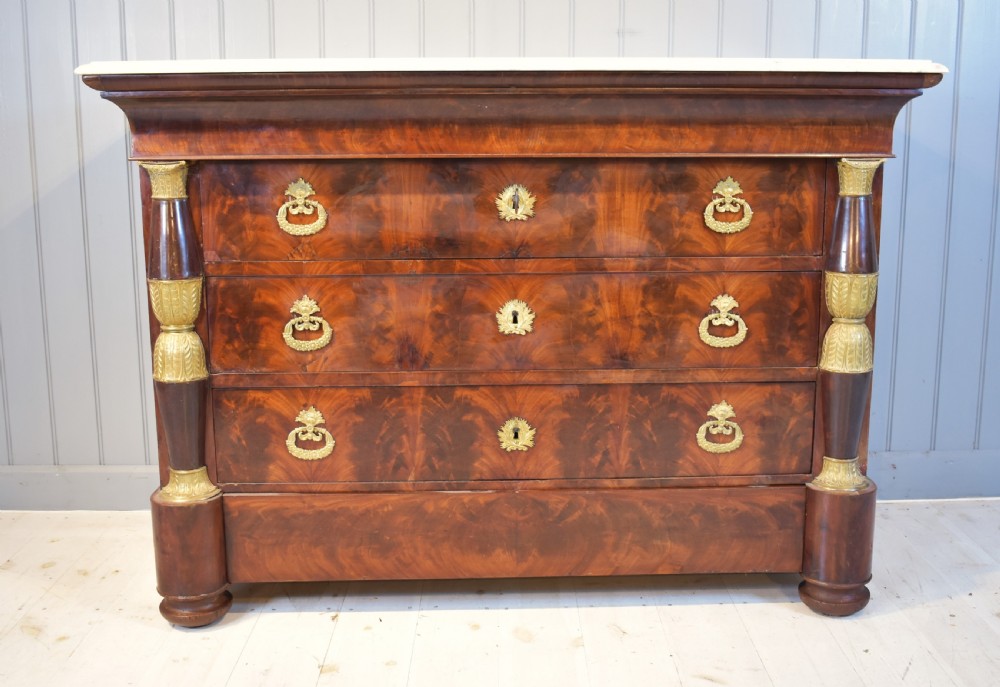 A 19th-century French Empire flame mahogany commode featuring a white marble top, three large drawers with gilt-bronze wreath handles, and flanking columns topped with ormolu accents.
