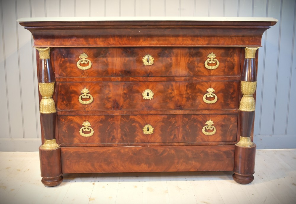 A 19th-century French Empire flame mahogany commode featuring a white marble top, three large drawers with gilt-bronze wreath handles, and flanking columns topped with ormolu accents. View II