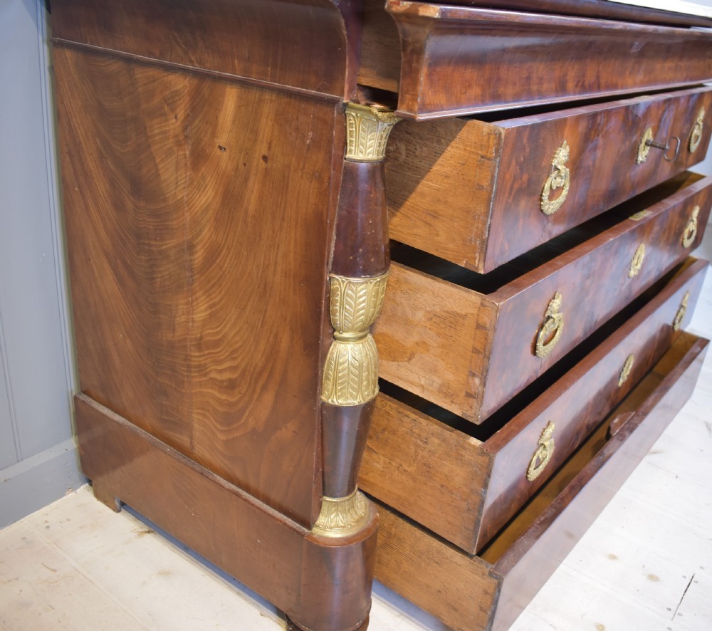 A 19th-century French Empire flame mahogany commode featuring a white marble top, three large drawers with gilt-bronze wreath handles, and flanking columns topped with ormolu accents. View VII
