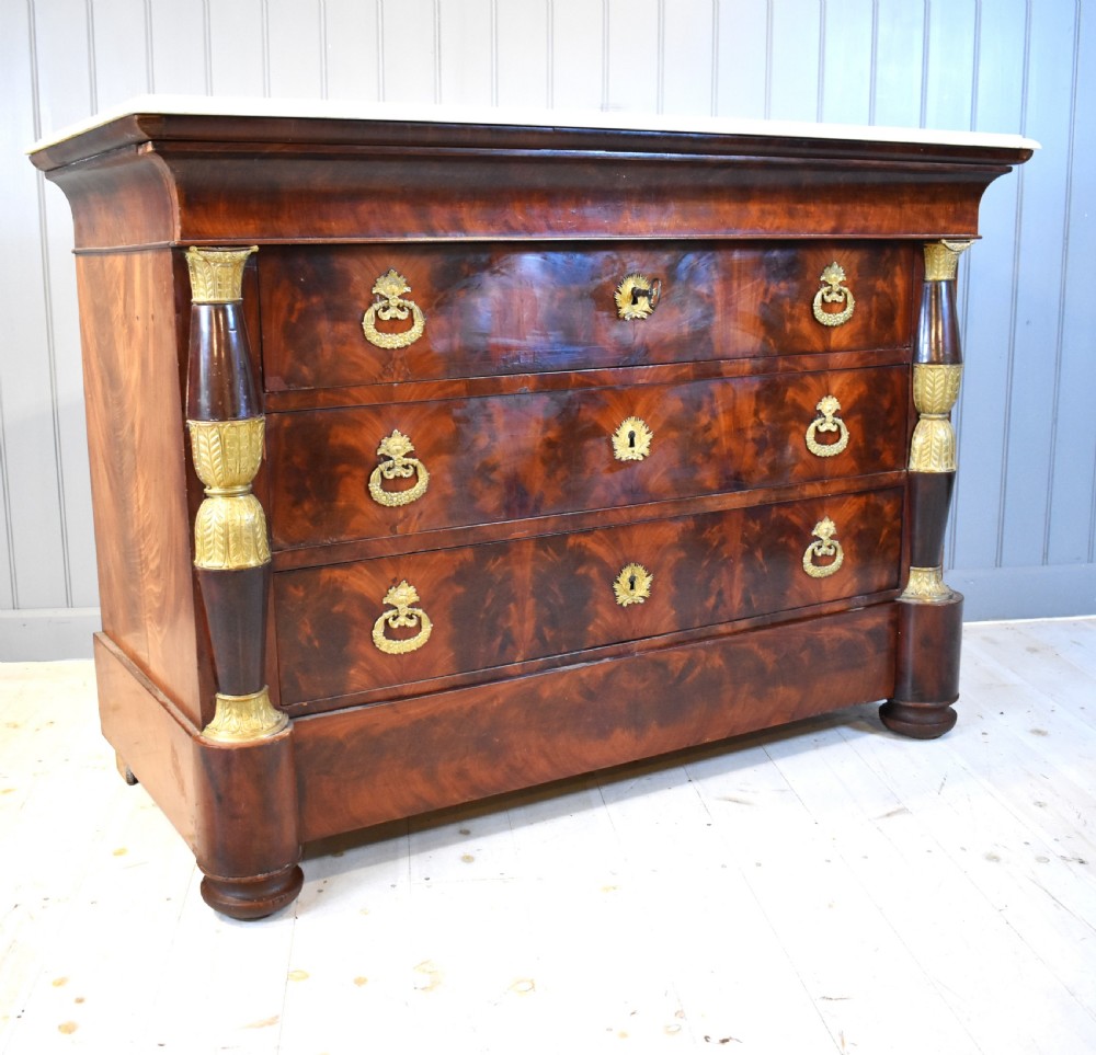 A 19th-century French Empire flame mahogany commode featuring a white marble top, three large drawers with gilt-bronze wreath handles, and flanking columns topped with ormolu accents. View XIV