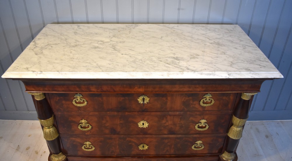 A 19th-century French Empire flame mahogany commode featuring a white marble top, three large drawers with gilt-bronze wreath handles, and flanking columns topped with ormolu accents. View XV