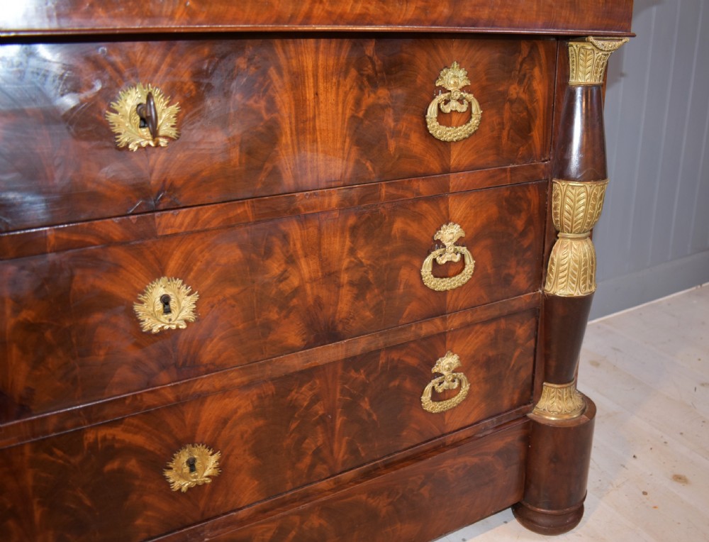 A 19th-century French Empire flame mahogany commode featuring a white marble top, three large drawers with gilt-bronze wreath handles, and flanking columns topped with ormolu accents. View XVI