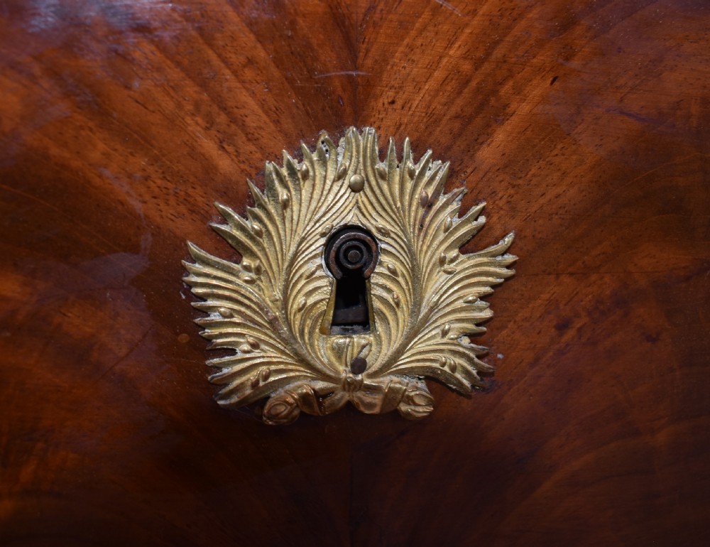 A 19th-century French Empire flame mahogany commode featuring a white marble top, three large drawers with gilt-bronze wreath handles, and flanking columns topped with ormolu accents. View XX