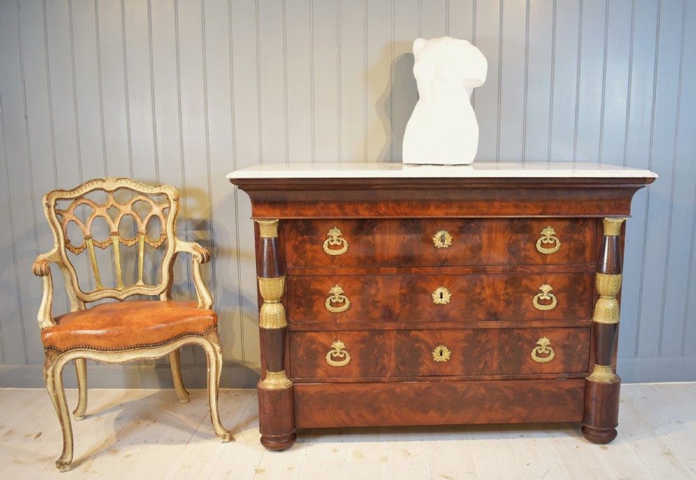 A 19th-century French Empire flame mahogany commode featuring a white marble top, three large drawers with gilt-bronze wreath handles, and flanking columns topped with ormolu accents. View XXII