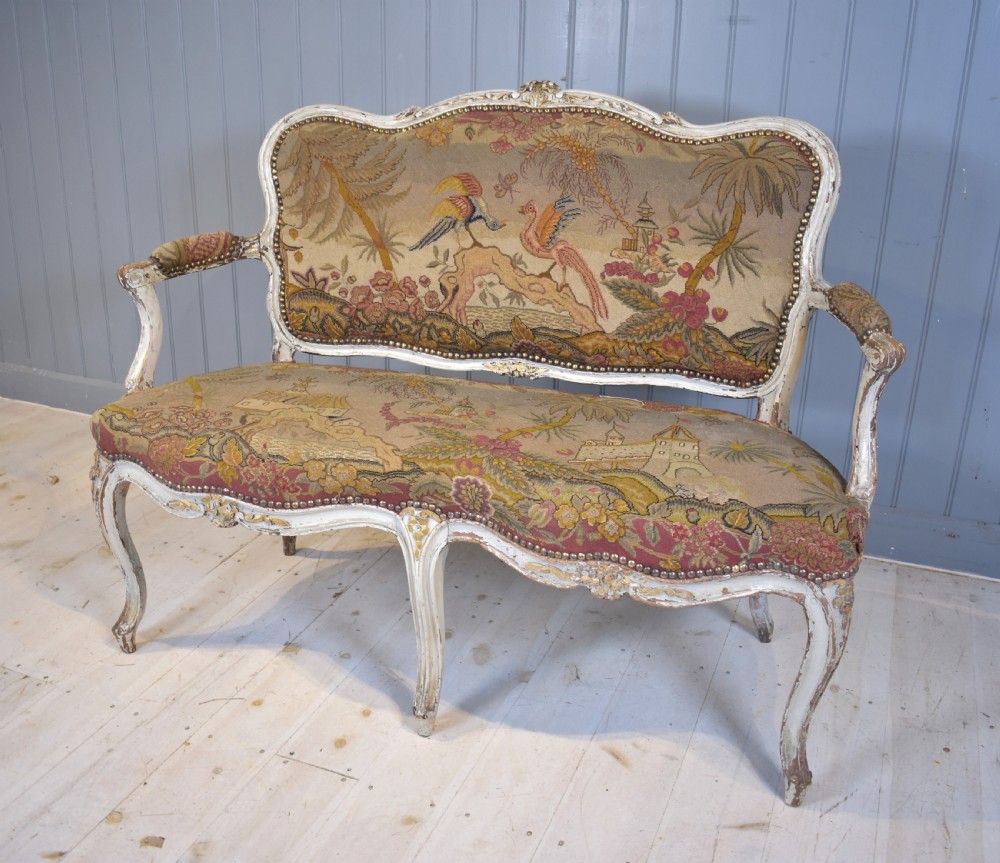 A French Louis XV style painted wooden settee with serpentine rails, cabriole legs, and woven tapestry upholstery featuring birds and palm trees.