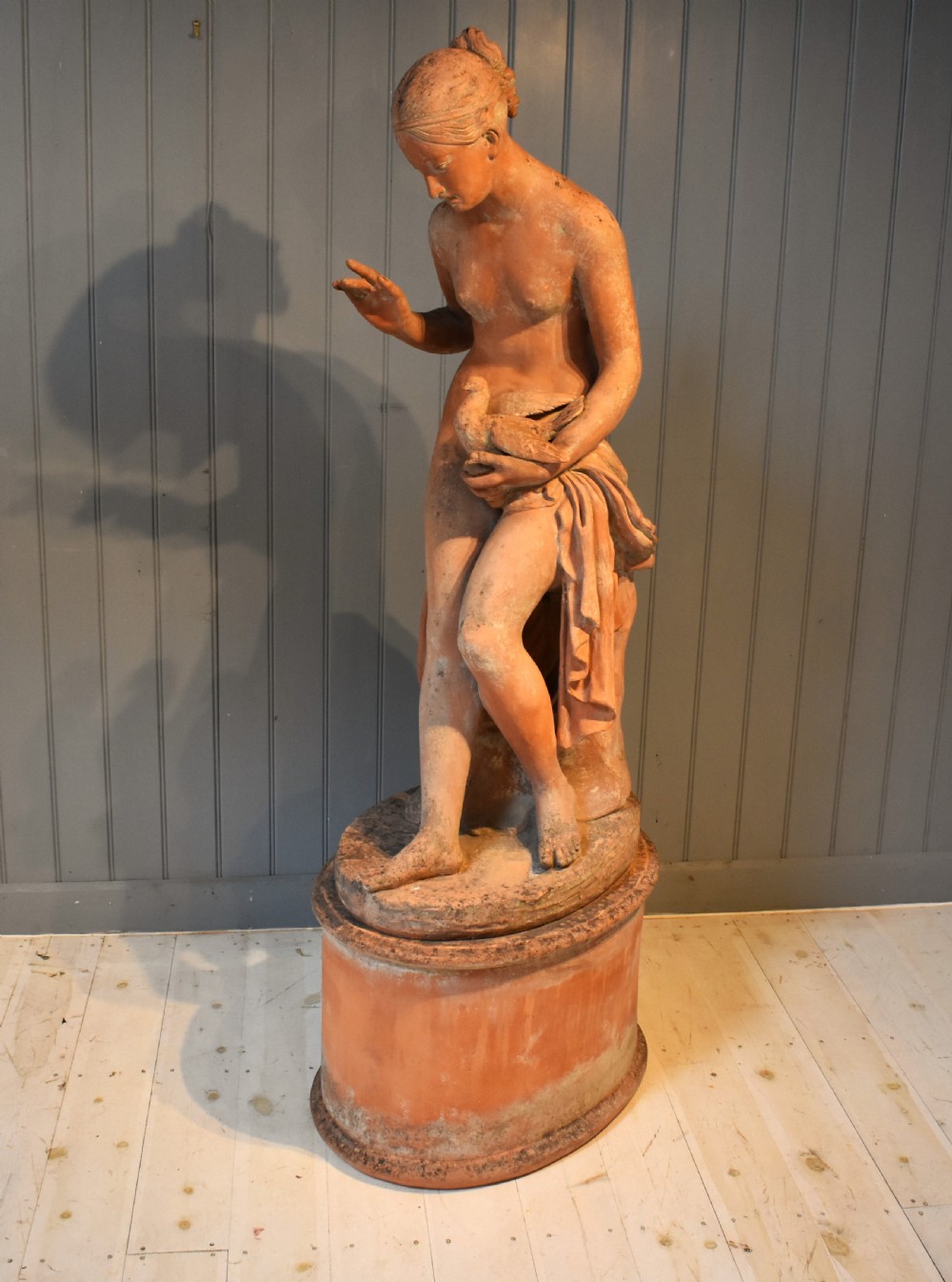 A classical terracotta sculpture of a seated draped female figure holding a bird, resting upon a matching oval terracotta pedestal.