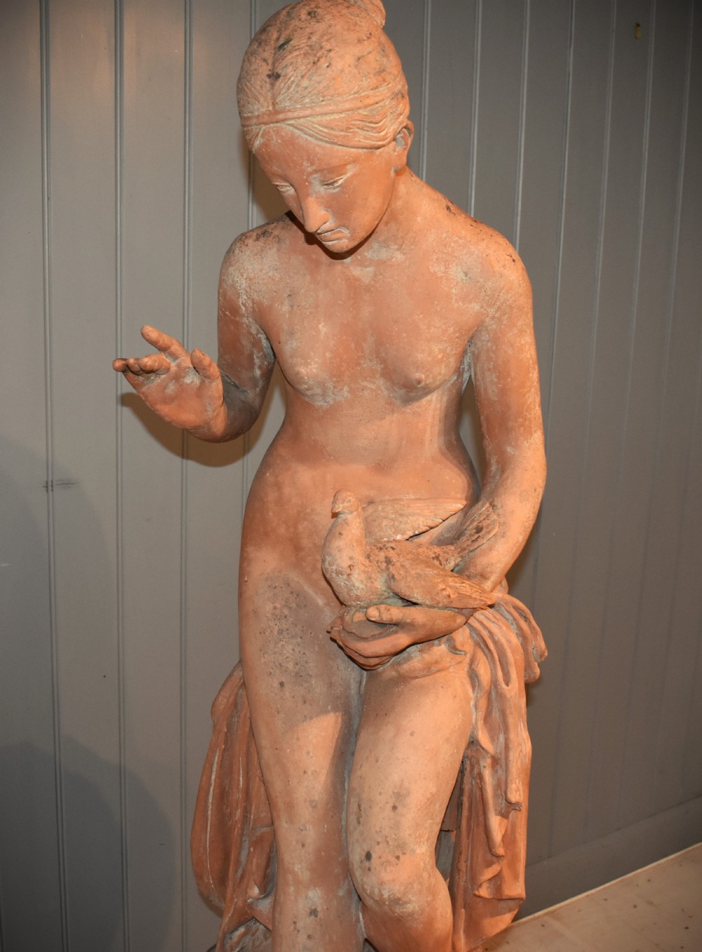 A classical terracotta sculpture of a seated draped female figure holding a bird, resting upon a matching oval terracotta pedestal. View II